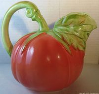 Front view of ceramic tomato-shaped pitcher, showing the red tomato body, green leaf spout, and curled handle.