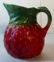 Side view of ceramic strawberry creamer showing detailed texture and color of red strawberry body and glossy green leaves forming handle and spout.