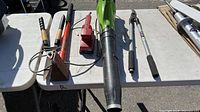 Top-down image showing Green Works leaf blower, Makita sander, log splitter axe, and manual plant trimmer and pruner laid out on a white folding table outdoors.