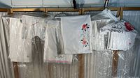Collection of six hand made kitchen linens hanging on hangers under plastic covers, some with floral embroidery.
