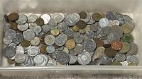 Top-down view of a plastic container filled with a large quantity of various vintage foreign coins and tokens in silver, bronze, and copper tones.