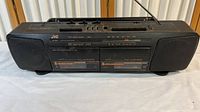 Front view of the black JVC RC-W210 boombox showing dual cassette decks, radio tuning scale, and speakers.
