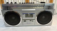 Front full view of the boombox showing dual large speakers, cassette deck with clear window, control knobs on top, and AM/FM radio dial.