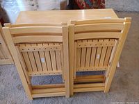 Folded wooden chairs showing the slatted wooden backrests and seats, in natural wood finish.