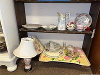 Shelf with various ceramic pitchers, floral figurine, porcelain dishes including plates, cups, and bowls, and a wooden bed tray with floral design. Lamp and shades at lower left corner.