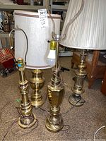 Four brass table lamps, two with pleated white fabric shades and two without shades. All lamps have brass bodies with various designs and visible wiring.
