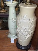 Two tall ceramic lamps with floral relief designs, one cream on black base and one celery green with no shades.