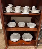 Shelf showing Royal Doulton Angelique china including stacked sets of tea cups, saucers, and an assortment of plates arranged on three shelves.