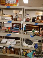 Two beige plastic shelving units filled with various household items in a store or auction setting. Shelves are tall and have multiple slats, with blue tape markers on them.