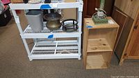 View of plastic shelving unit with three levels and unfinished two-compartment wooden shelf box next to it, both on floor indoors.