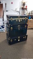 Three vintage dark green steamer trunks stacked vertically showing front and side views, brass hardware, locks and leather handles, one missing a handle, scuffed/scratched finish