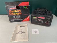 Photo showing the battery charger unit, original box, and printed manual laid out on a green background surface.