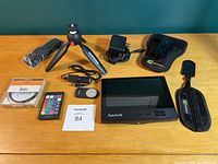 Photo showing full camera accessories kit laid out on a wooden table against a green wall, including tripod, monitor, filter, charger, remote, cables, and straps.