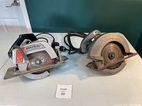 Two circular saws placed side by side on a white table with green wall background. One is a Black & Decker 7.25 inch saw and the other is a Skilsaw 8.25 inch saw. Both have cords and show rust and wear.