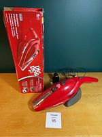 Red Dirt Devil Scorpion handheld vacuum cleaner with original box, charging base on wooden surface against green background.