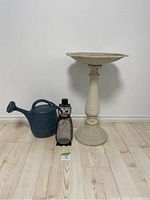 Wide view showing all three items: metal penguin bird feeder, blue watering can, and cream metal bird bath with pedestal.