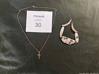 Photo showing gold cross necklace with delicate chain and multi-coloured linked metal bracelet laid on black fabric background with a maxsold sign.