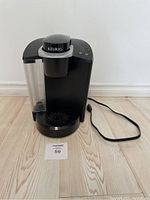 Front view of black Keurig K40 coffee maker with transparent water reservoir on left side, power cord extended on floor.