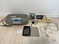 Sony boombox-style radio, HP tablet with folio case, TomTom GPS device, power strip, and charging cables on wooden floor.