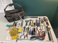 Photo showing the tool pouch with the metal handle and light, along with all hand tools laid out on a white table, including gloves, hammer, scrapers, files, wrenches, chisels, calipers, pliers, saw and other tools.