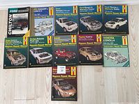 Photo showing eleven Haynes auto repair manuals arranged in two rows on a wooden floor. Covers depict various vehicle images and Haynes branding.