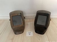 Both welding helmets shown side by side on floor, one rusted metallic, one black.