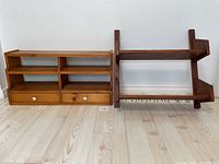 Photo showing both wooden shelf units side by side on floor against wall. The lighter wood unit is on the left with two drawers and open shelves. The darker wood unit is on the right with hooks under the bottom shelf.