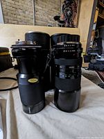 Both zoom lenses and their accompanying hard cases side by side on a cloth surface. Lenses are black with aperture and focus rings visible.
