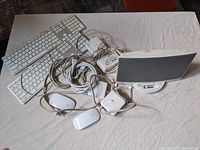 Two Apple wired keyboards, two Apple Magic Mouse devices, three Apple MagSafe chargers, and one Bose charging dock speaker with docking port, all on a beige surface.