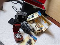 Vintage cameras with light meter and accessories arranged on wood surface near hard carrying case.