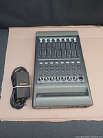Front view of Mackie Logic Control XT with eight channel faders and control buttons, and the accompanying power adapter coiled beside it.