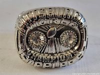Front view of replica Pittsburgh Steelers World Champions football ring showing raised text, Lombardi Trophy symbol and two clear stones.