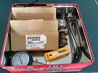 Full lot inside red metal container showing ignition coils in cardboard boxes, NGK spark plug box, towing hitch ball, pressure gauge, and automotive tools.