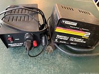Photo of two battery chargers, including a Modlastar MC2 charger and a regulated 12 Volt power supply, placed on a green surface with power cords attached.