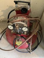 Front angled view of red Air-Pac piston air compressor showing pressure gauges, air hose, and power cord.