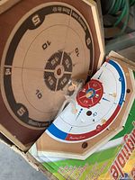 Wooden crokinole board showing the scoring rings and hole in middle, placed inside original cardboard box with colorful graphics, wooden game pieces visible.