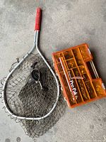Fishing net with metal frame handle and orange tackle box on concrete floor