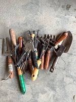 Overall view of gardening tools including trowels, cultivators, forks, and pruning shears with wooden and plastic handles laid out on a concrete floor
