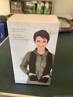 Box front showing woman wearing the black heated cable knit scarf with hand pockets, product name and description.