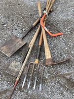 Photo showing collection of lawn care tools laid on gravel surface including a large and small shovel, pitchfork, pickaxe and bow saw.