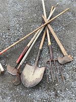 Top view of six garden tools laid on gravel: broom, hand cultivator, shovel, pitchfork, hoe, and a tool with flat blade.
