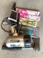 Photo of small hatchet, GPS device, pencils, drawing compass, and large flashlight arranged on floor