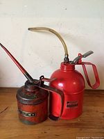 Two vintage metal oil cans placed on a wooden surface against a plain white wall; the smaller is rusted with a long thin spout and Pesco label, the larger is bright red with clear flexible tube and lever handle.