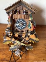 Front view of the old wooden cuckoo clock showing detailed carvings of birds and rifles, the clock face, and dancing figurines at the top.
