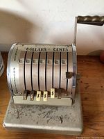 Front view of Paymaster mechanical money machine showing numeric dials, handle, and dollar sign buttons; machine surface is dusty and worn.