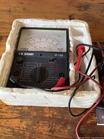 Front view of the A.W. Sperry SP-1708 analog multimeter showing dial, probe wires, and needle display, resting in white foam packaging.