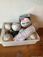 Photo showing shallow beige container with five baseballs, one boxed Blue Dot softball, assorted small colored sports balls, and a white mesh item.