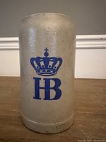 Full front view of the 1 litre German beer stein showing the gray textured stoneware and blue HB crown logo design.