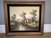 Full view of the framed oil painting showing a pastoral landscape with a river and a house, surrounded by trees.