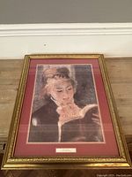 Framed print of a woman reading a book by Renoir, showing full framed artwork and matting against a wooden floor background.